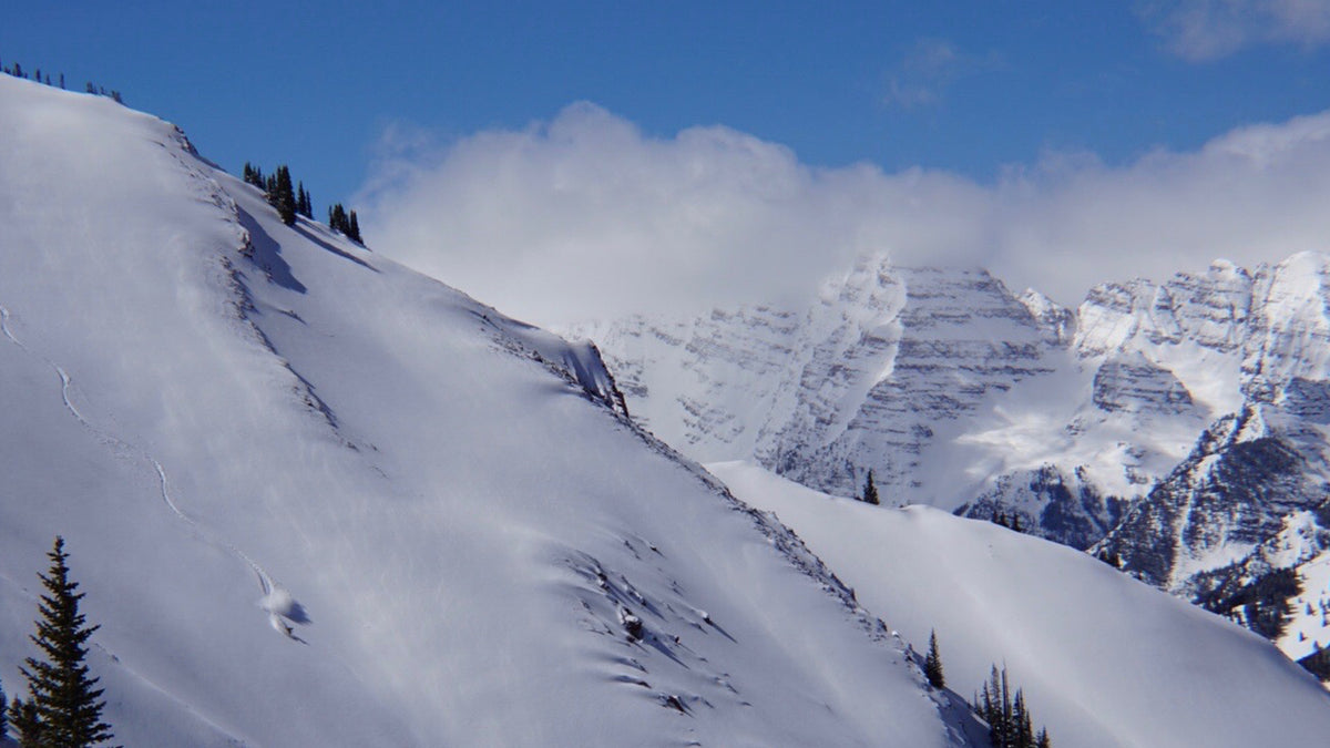 Snow-covered mountain slopes under a blue sky with clouds, featuring evergreen trees and rugged rocky peaks in the background.